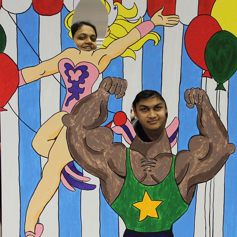 Photo: Nupura Kale and Jay Ramanuj smile through trapeze and strongman cutouts at the Neumann Library Open House Carnival. Photo by The Signal reporter Cindy Brady.