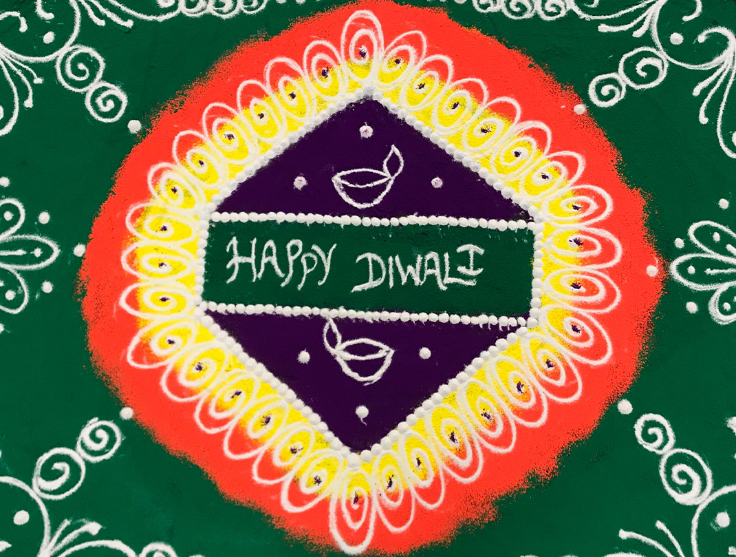 This photo features sand art done for the Diwali celebration. It starts with green the goes to orange, yellow, purple, then back to green. The center features the words "Happy Diwali" in the center.