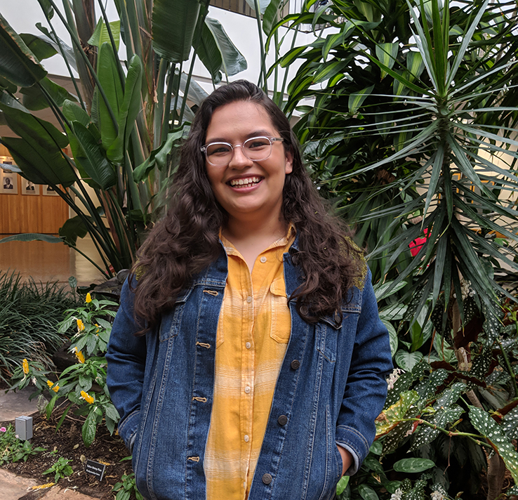 Natalie Garcia wears a yellow shirt and jean jacket, posing in front of the inside garden in the Bayou Building at UHCL.
