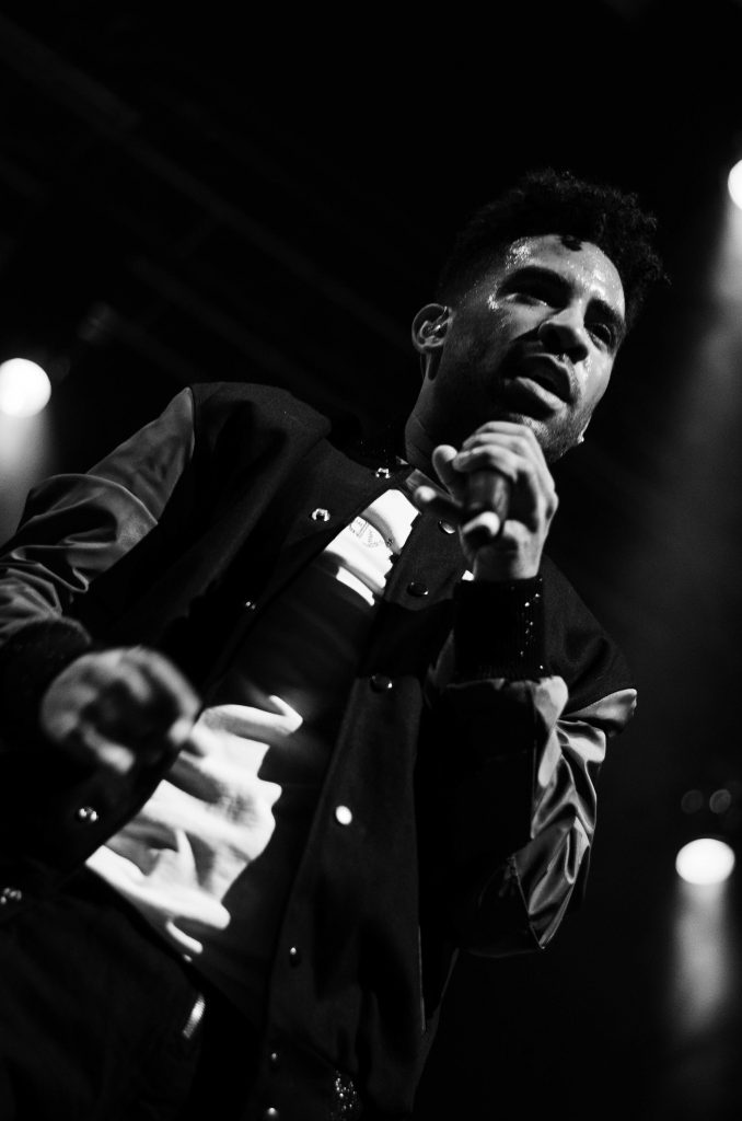PHOTO: Black and white photo of singer Kyle performing one of his songs. He is wearing a sports jacket and performing with a microphone. Photo courtesy of Shalnora Worlds.