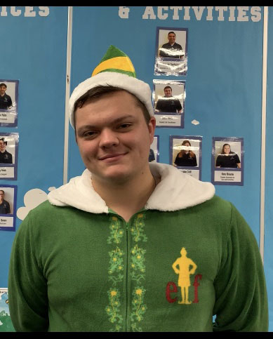 PHOTO: Justin Murphy dressed in a green onesie and a green and yellow Santa hat in front of a blue bulletin board. Photo by Signal Co-Managing Editor Troylon Griffin II.