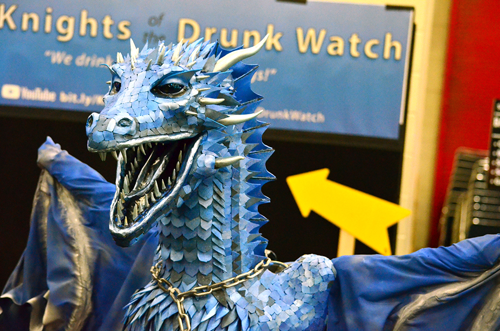 PHOTO: A mixed-media statue of a blue dragon made out of metal scales in various shades of blue and silver stares off camera with its mouth open as if in a roar and its wings spread. A sign that reads “Knights of the Drunk Watch” can be seen in the background. Photo by The Signal reporter Jennifer Martinez.