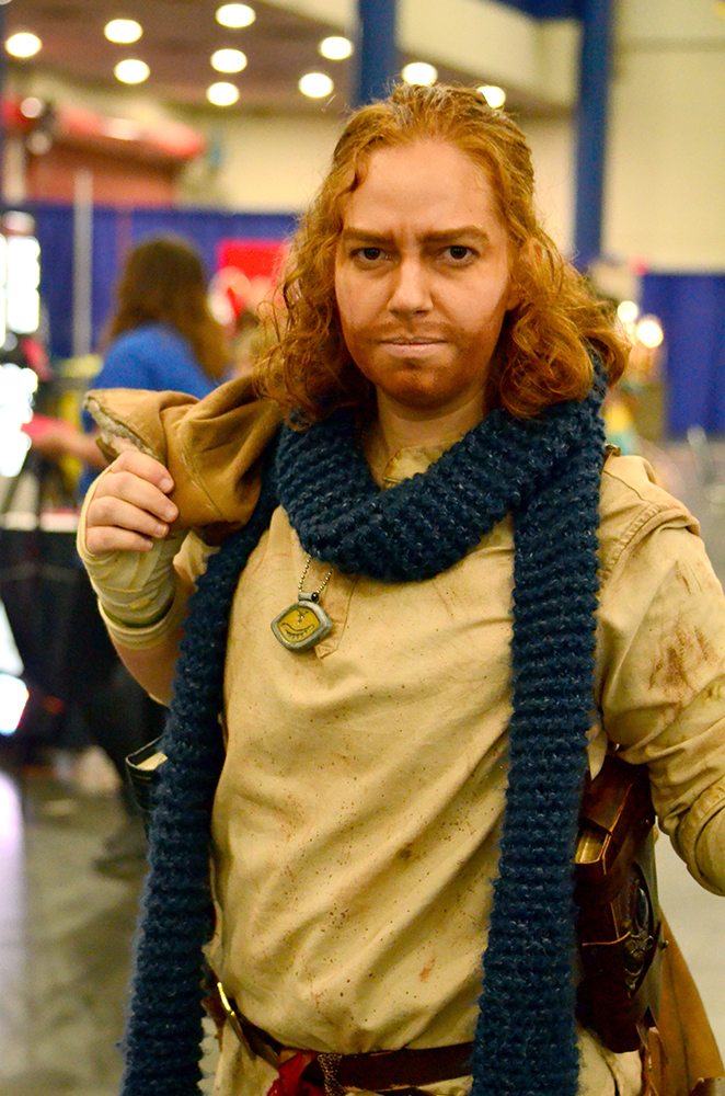 PHOTO: A woman with shoulder-length red, curly hair and a makeup red beard painted on her face to make her look like the male character she is cosplaying as poses in the costume designed after Caleb Widogast from Critical Role Season 2. The costume includes a simple tan shirt, a blue scarf, bandages wrapped around the hands, a long brown leather jacket with fur fleece lining and leather holsters carrying books on each side of the waist. Photo by The Signal reporter Jennifer Martinez.