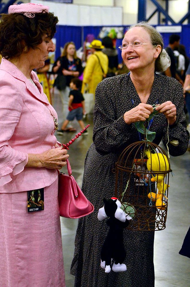 Two older women cosplayers talk to each other in costume. The woman on the left is wearing an all pink skirt and blazer suit, wearing a small pink hat and carrying a matching pink purse and wand set. The woman on the right is wearing a simple black dress with polka dots and is holding a birdcage with a stuffed Tweety toy inside it and a stuffed Sylvester toy hanging from the outside, as if it is trying to get in to the Tweety doll. Photo by The Signal reporter Jennifer Martinez.