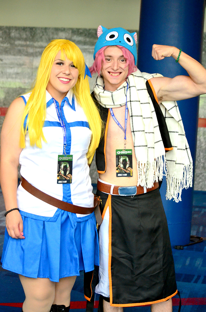 PHOTO: A young woman and man in costume pose next to each other. The woman stands on the left. She has a long blond wig with a blue bow and the right side of her head, is wearing a sleeveless white shirt with blue trim, a matching blue skirt and a brown leather belt. The man is on the right. He is wearing a blue cat knit cap, a pink wig, an oversized white scarf with thin black stripes, an open black vest with orange trim, no shirt and a black skirt with orange trim. Photo by The Signal reporter Jennifer Martinez.