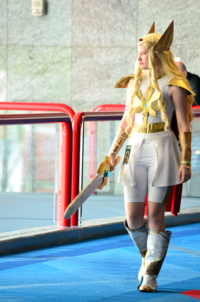 PHOTO: A young woman dressed as She-Ra from the Netflix original “She-Ra and the Princesses of Power” walks towards the camera with he face pointed off-camera. She is wearing a long blonde wig, a gold headband with wing-like protrusions on either side of the head, a white outfit with gold highlight, gold shoulder armor, gold wrist guards and gold and white boots. She is carrying a fake sword with a large golden handle that has a blue gem in the center.