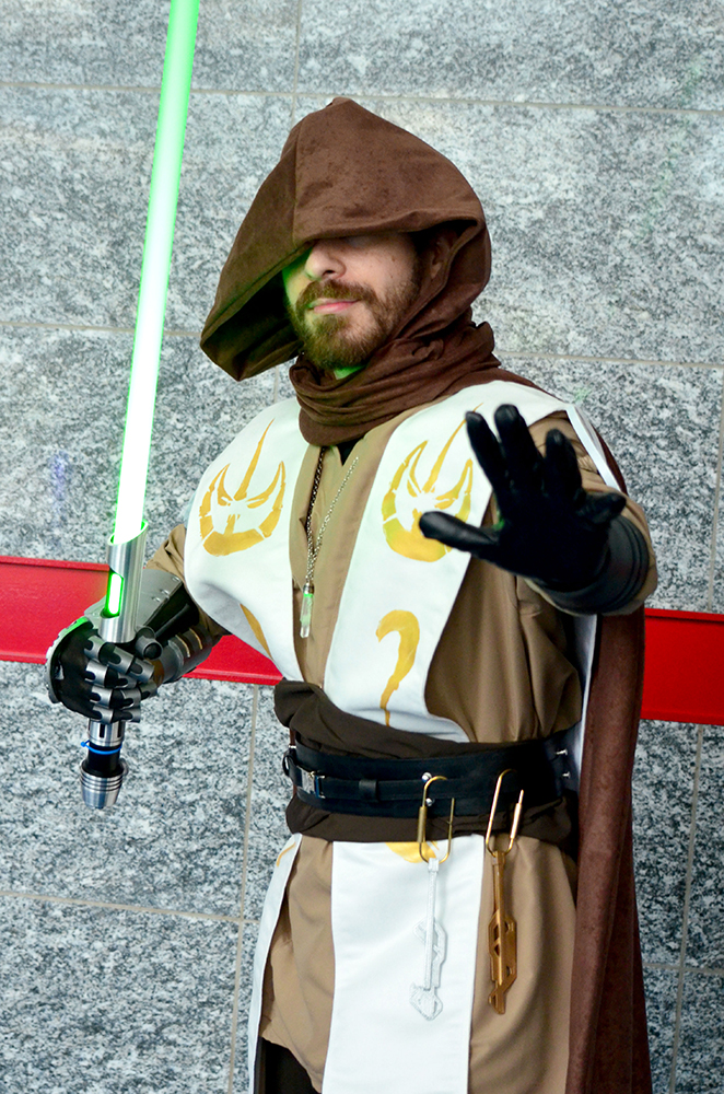 PHOTO: A man dressed in brown robes with a white and gold mantle over top and a dark brown hood covering the top half of his face poses while holding a green lightsaber. Photo by The Signal reporter Jennifer Martinez.