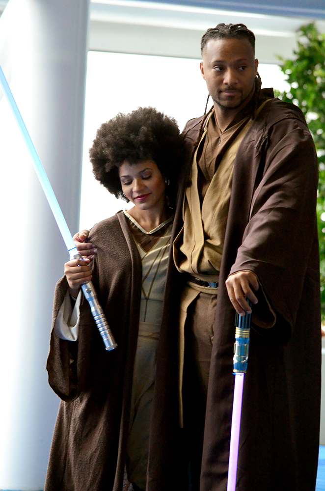 PHOTO: A woman and a man dressed as Jedi from Star Wars lean on each other. The woman is standing on the left, the man on the right. They are both wearing simple brown robes with tan undershirts and pants. The woman is carrying a blue lightsaber, the man is carrying a purple one. Photo by The Signal reporter Jennifer Martinez.