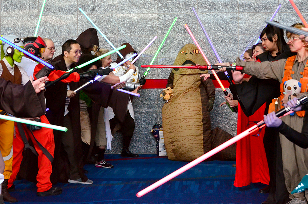 PHOTO: A crowd of cosplayers carrying lightsabers of various colors face each other in challenge while a person in a Jabba the Hutt costume stands between them with a confused look on their face. Photo by The Signal reporter Jennifer Martinez.