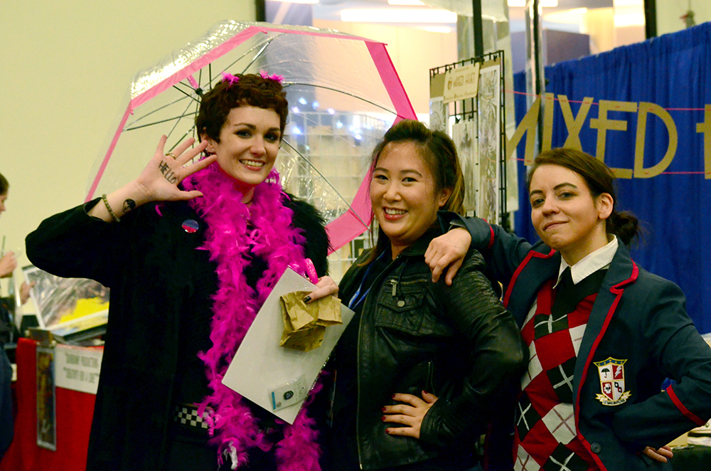 PHOTO: Three people stand next to each other. The person to the far left is holding up their hand that has “Hello” written on it, while balancing a clear umbrella with a pink trim in the other. They are wearing a pink feather boa and a Bisexual Pride button. The rest of their outfit is black. The person in the middle is wearing a black leather jacket. The person on the far right is wearing a preparatory school uniform that includes a blue blazer with red trim, a white undershirt, a blue, grey and red checkered sweater vest and a school logo on the blazer. Photo by The Signal reporter Jennifer Martinez.