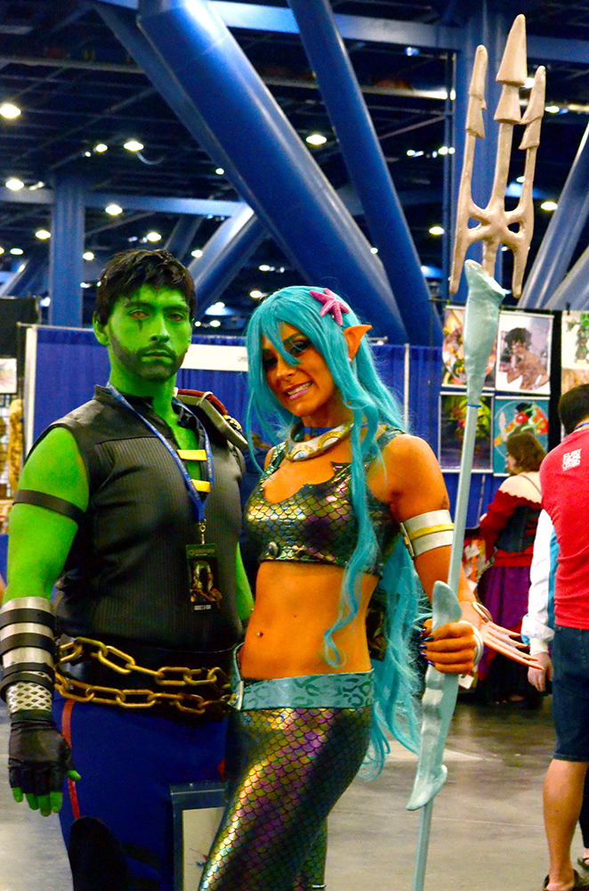 PHOTO: A man and a woman dressed as characters from the show Reboot pose together. The man is on the left, the woman on the right. The man has green painted skin and is wearing a black sleeveless shirt, blue pants, and a large gold chain belt. The woman has orange painted skin, a long teal wig, and is wearing holographic fish scale print bodice and pants. She is also holding a trident. Photo by The Signal reporter Jennifer Martinez.