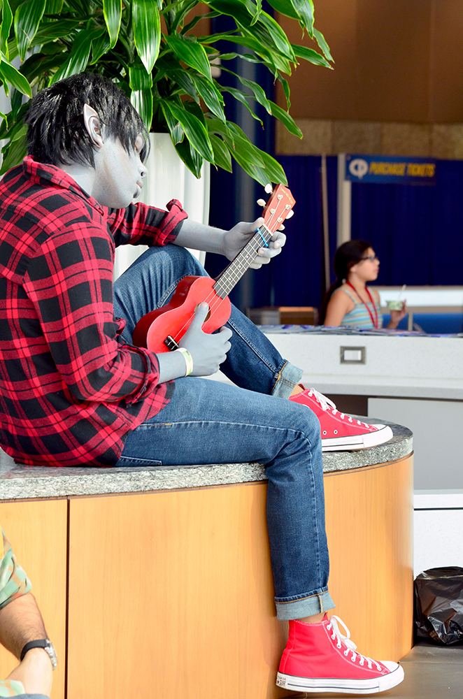 PHOTO: A young man with grey painted skin, a red and black plaid shirt, jeans and red high-tops calmly strums a red ukulele while sitting in a relaxed pose on top of a bench. Photo by The Signal reporter Jennifer Martinez.