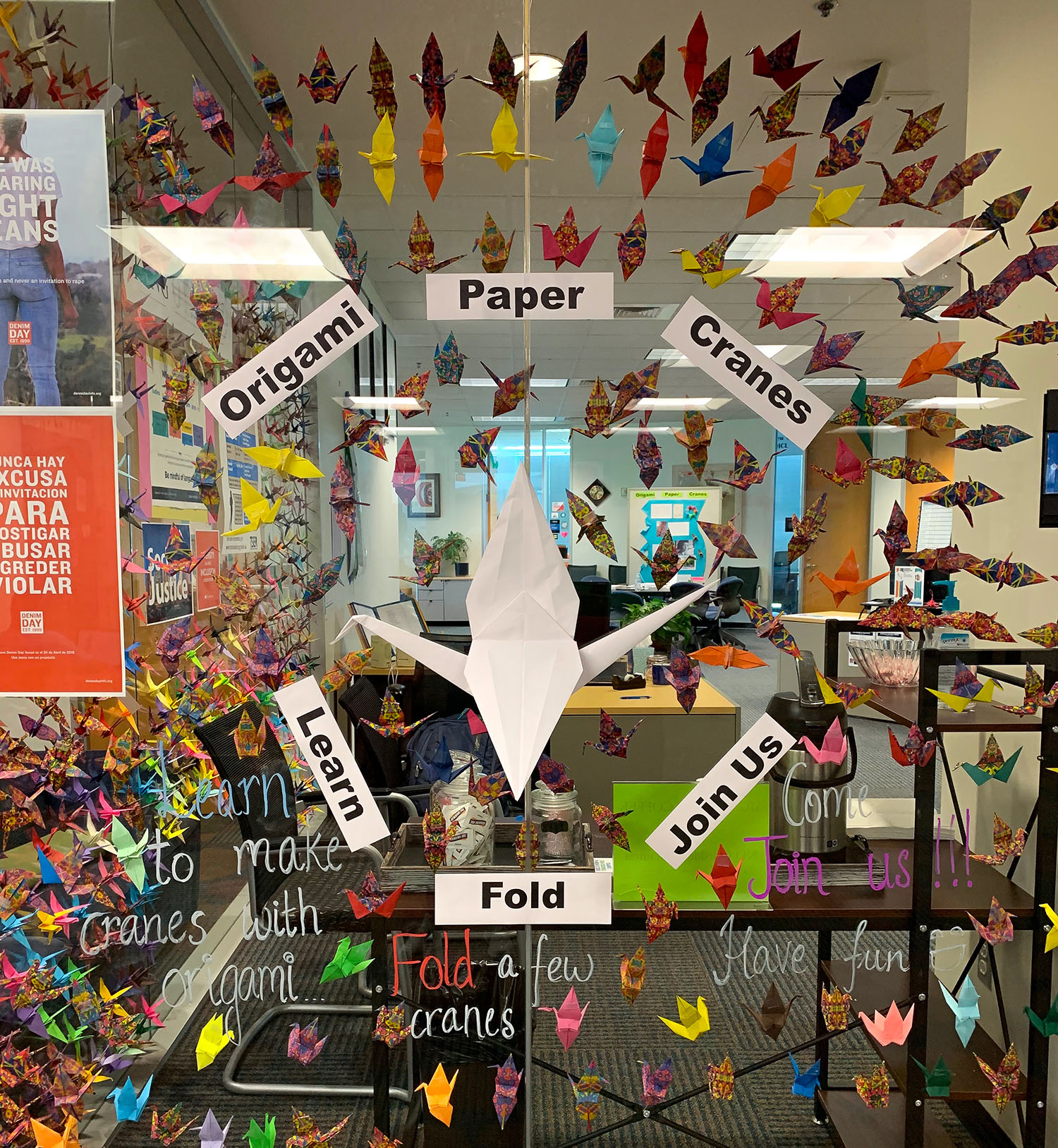 PHOTO: Photo of window filled with an array of different colored, origami paper cranes. Text stating learn, fold and join us.