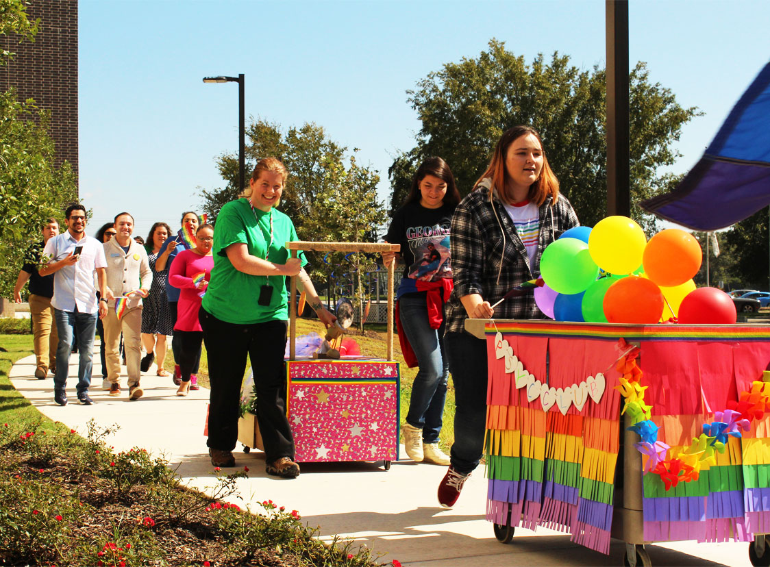 PHOTO: Image depicts a line of people walking down a sidewalk holding colorful posters and floats for the UHCL Pride Parade.Photo by The Signal reporter Arturo Guerra.
