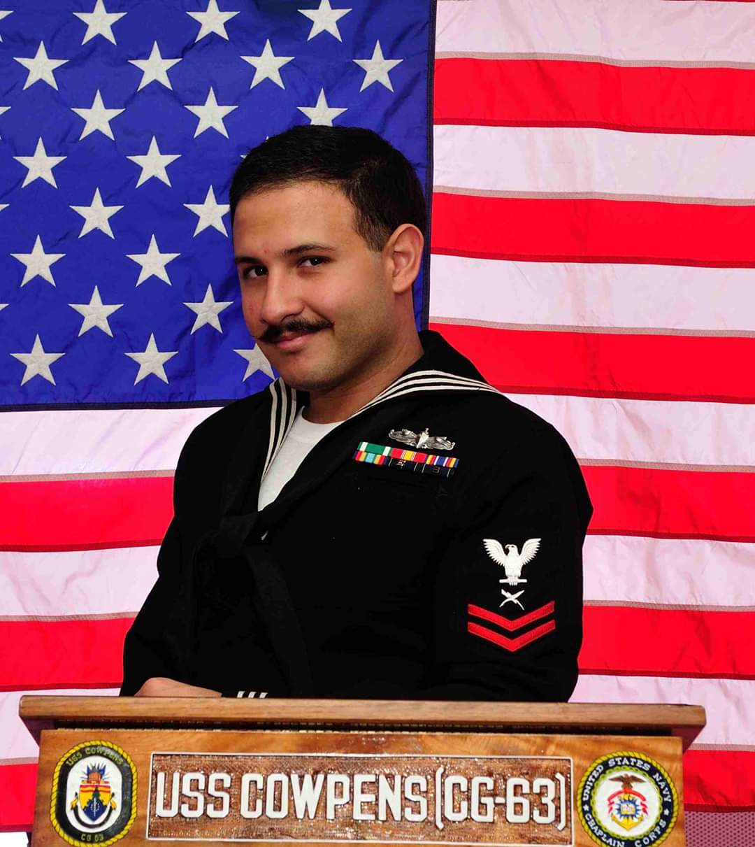 PHOTO: Photograph of veteran student Arturo Guerra in uniform while overseas. In the background of the picture you can see the American flag as well as Arturo Guerra leaning on a podium that reads USS COWPENS [CG-63]. Photo courtesy of Arturo Guerra.