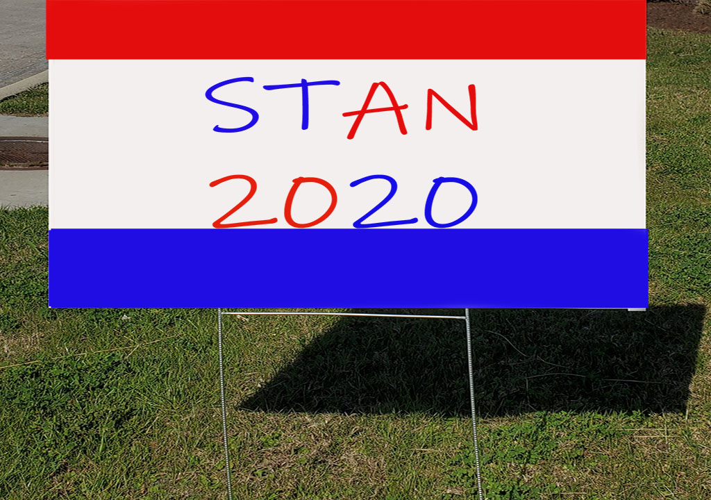 ILLUSTRATION: Red, white, and blue political sign that reads "STAN 2020" in red and blue letters in a grassy area on the side of the road.
