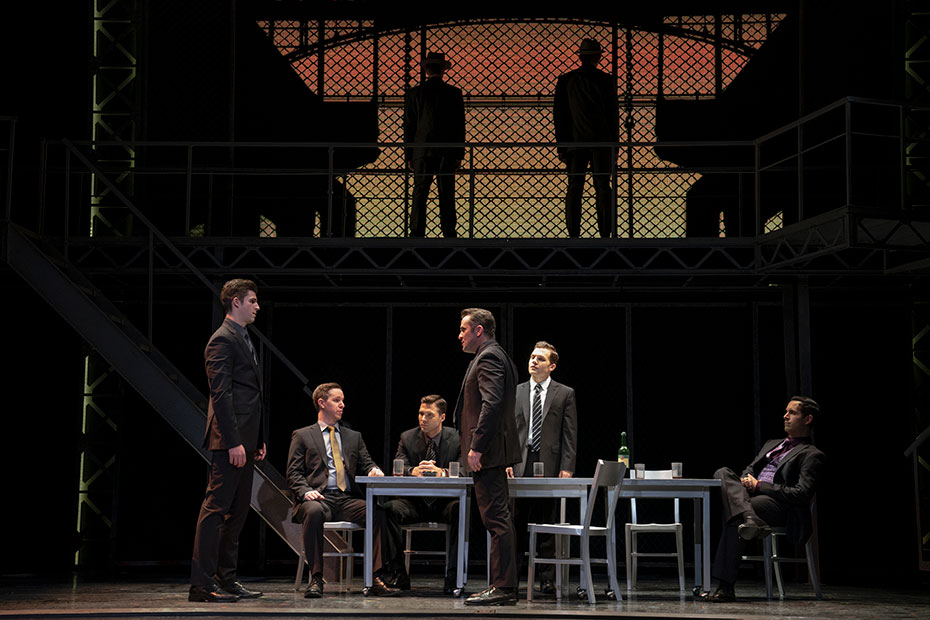PHOTO: The Four Seasons, the loan shark and Angelo DeCarlo stand off at a table while two thugs are silhouetted above them upstage. Left to right: Eric Chambliss, Kevin Patrick Martin, Michael Milton, Corey Greenan, Jon Hacker and Andrés Acosta. Photo by Joan Marcus and courtesy of Jersey Boys on Tour.