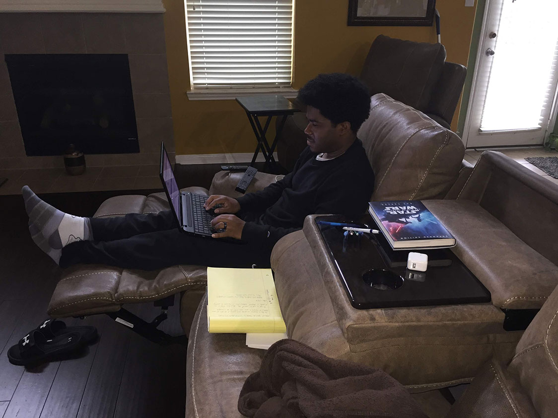 PHOTO: Image depicts young adult male on a couch typing on his laptop. Next to him is a book, notepad and pens. Photo by The Signal managing editor Troylon Griffin II.