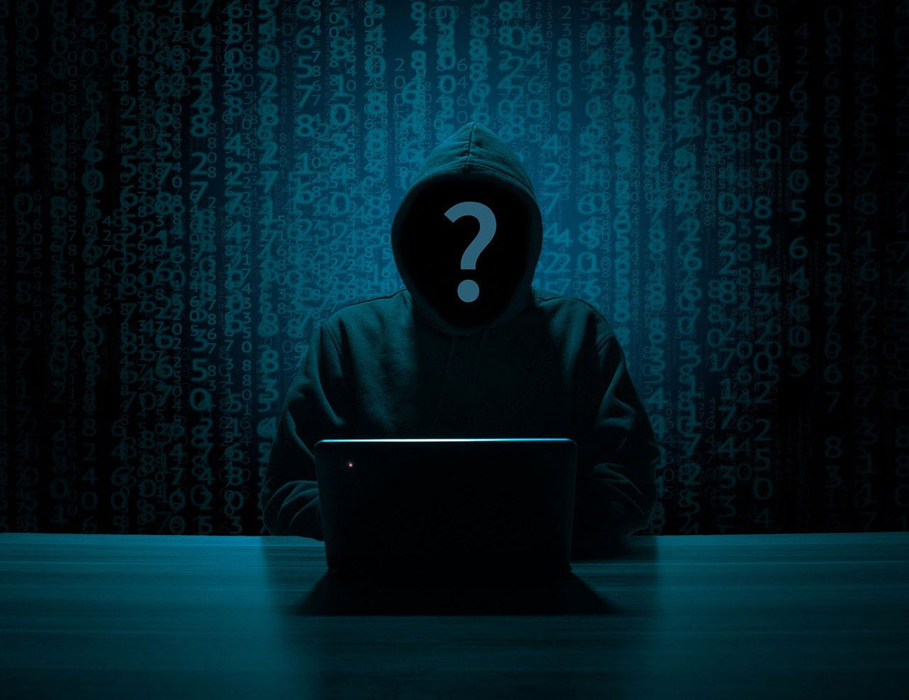 The photo shows a silhouette of a person in a black hoodie in front of a laptop. The background shows coding in a blue color showing the person is up to no good. The photo is a representation of a person violating another persons security on video conference calls.