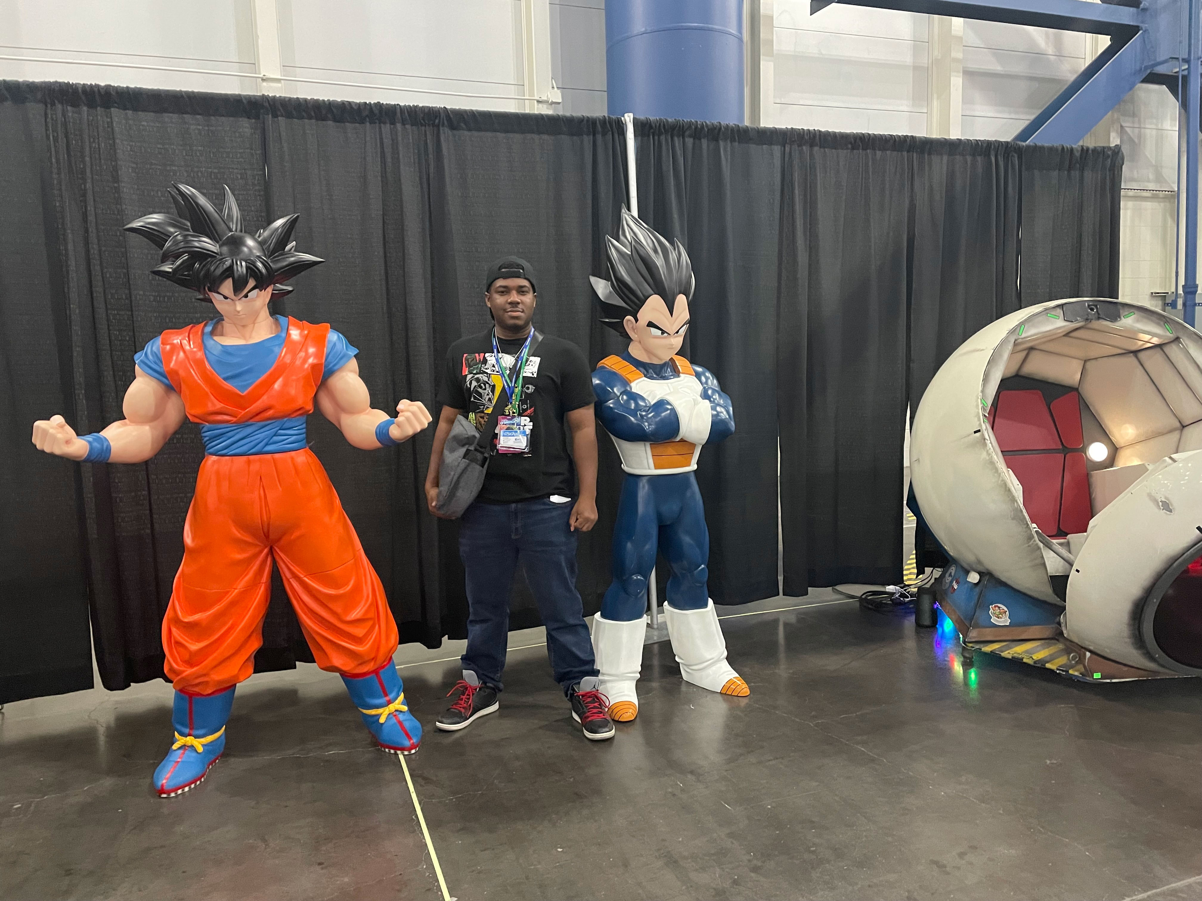 PHOTO: Image depicts boy with bag between two life sized statues of Dragon Ball characters Goku and Vegeta. They stand in front of black curtains next to a replica of a fictional space pod. Photo by The Signal Managing Editor of Content and Operations Troylon Griffin II.