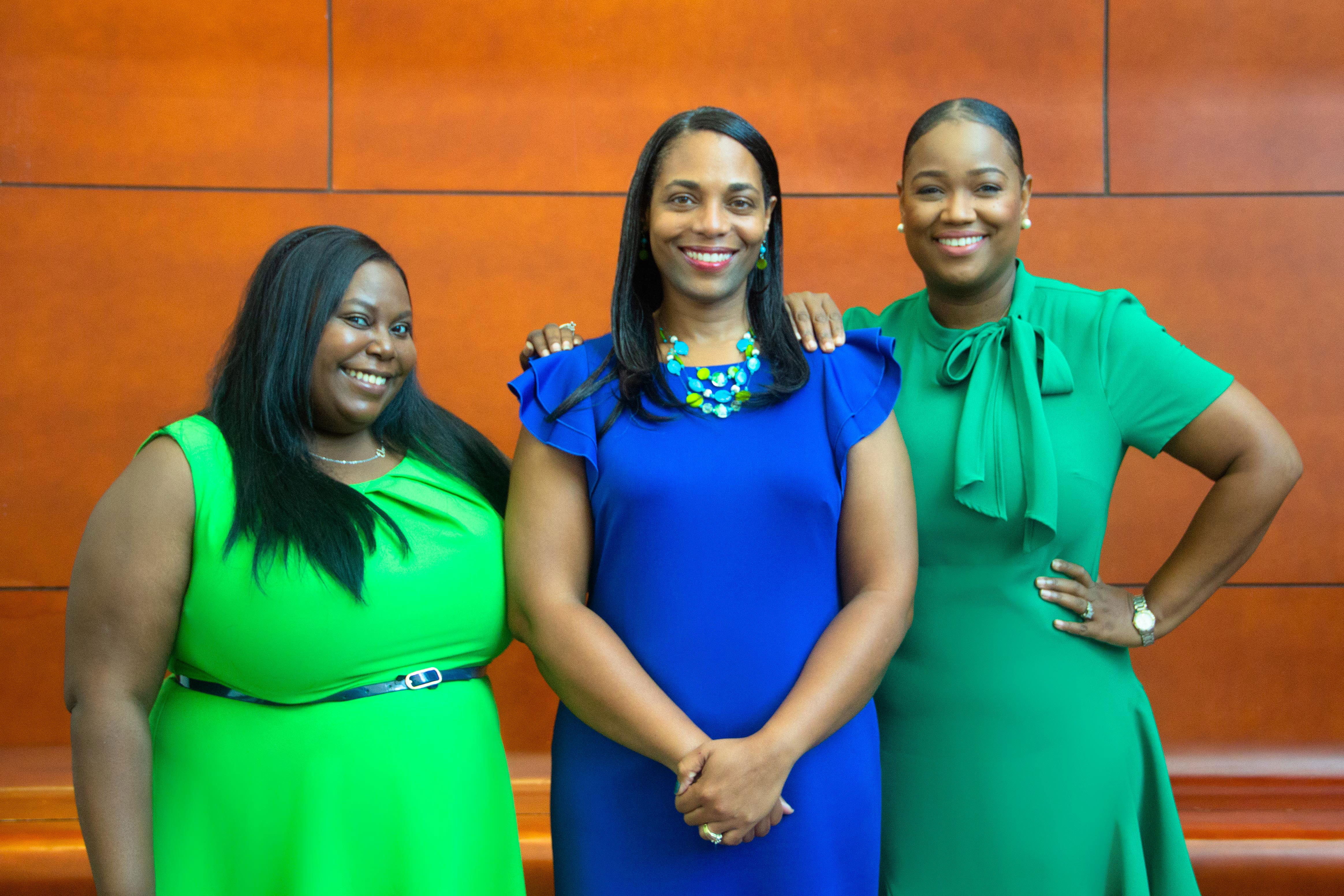 PHOTO: Photo consists of three women who are the staff of UHCL’s Office of Student Advocacy. The staff consists of the following (from left to right): Ebony Brunn, who is in a bright green dress, Kristi Randolph Simon, who is in a blue dress and LaToya Mills, who is in a darker green dress. All three are smiling with a brown wall behind them. Photo Courtesy of Ebony Brunn.