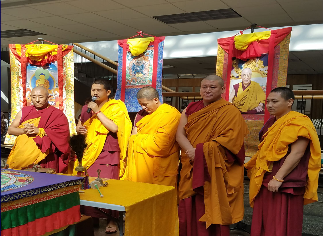 PHOTO: Image depicts five Tibetan Buddhist monks amidst a Buddhist backdrop. One is holding a microphone. All monks are in red and yellow robes. Photo by The Signal reporter Erica Bernal.