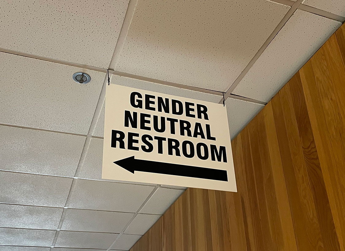 PHOTO: Image depicts white sign that says "GENDER NEUTRAL BATHROOM" with an arrow underneath the words pointing left. The sign hangs from a ceiling. Photo by The Signal Managing Editor of Content & Operations Troylon Griffin II.