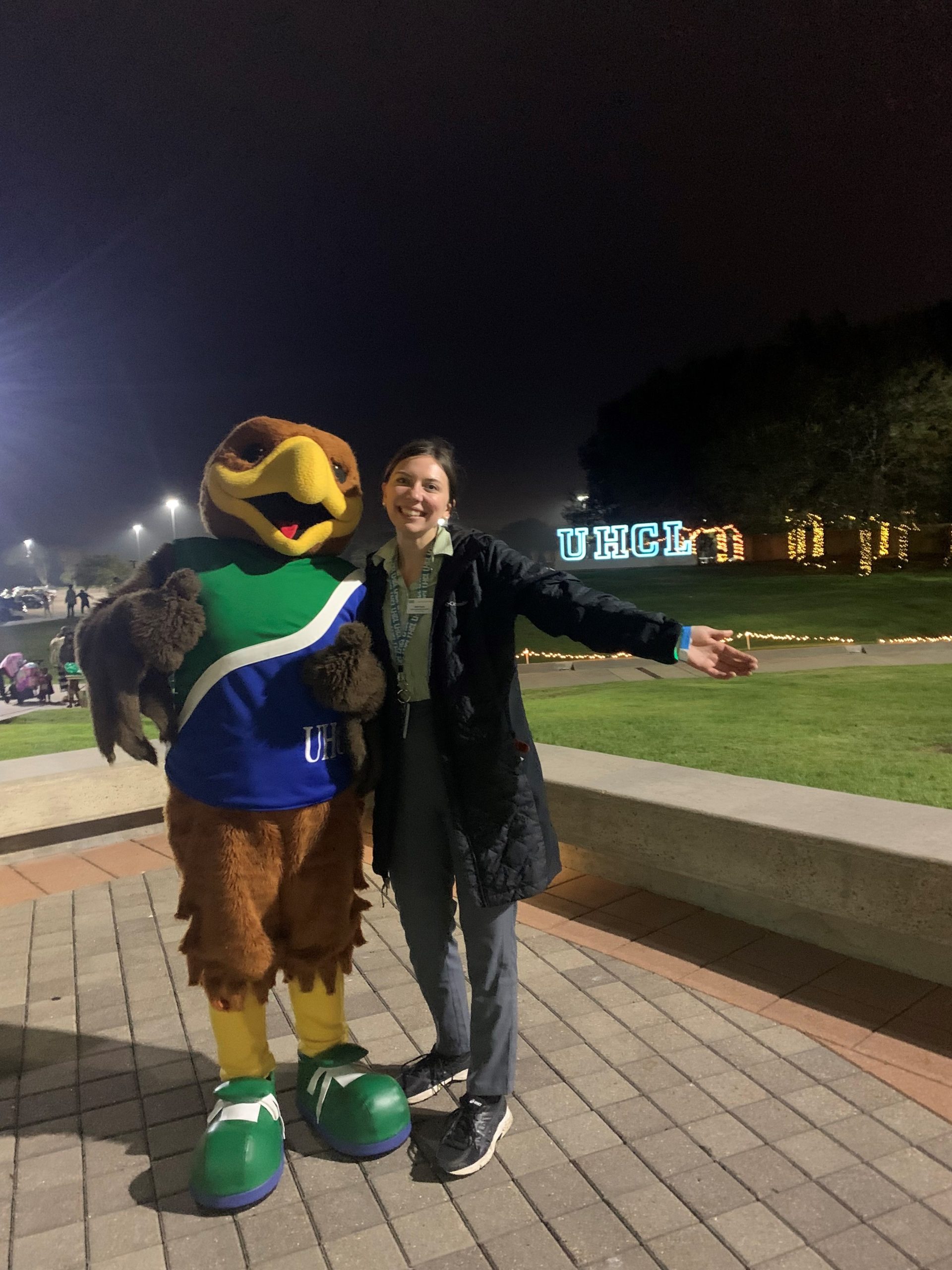 IMAGE: A photograph of Shelby Kuepker (right) standing next to UHCL Mascot Hunter the Hawk (left). In the background is the UHCL letters hill, lit up at night alongside a few trees with string lights wrapped around them.