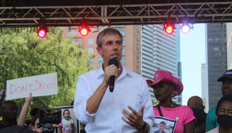 PHOTO: Beto O'Rourke speaks on stage. Behind him stands a person in a pink dress and cowboy hat and protester holding a sign. Photograph by The Signal Managing Editor Cesar Cardenas.