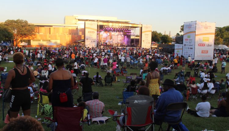 A did shot of the main stage from the center back of the crowd. People are on their feet as Sheila E. performs. On the right you can see the scaffolding where the production team did technical work. White banners flank the stage with a list of sponsors from h-e-b to reliant. The sun is setting on the stage as flashing purple lights beam out from the top. Photograph by The Signal reporter Jared Cadore.