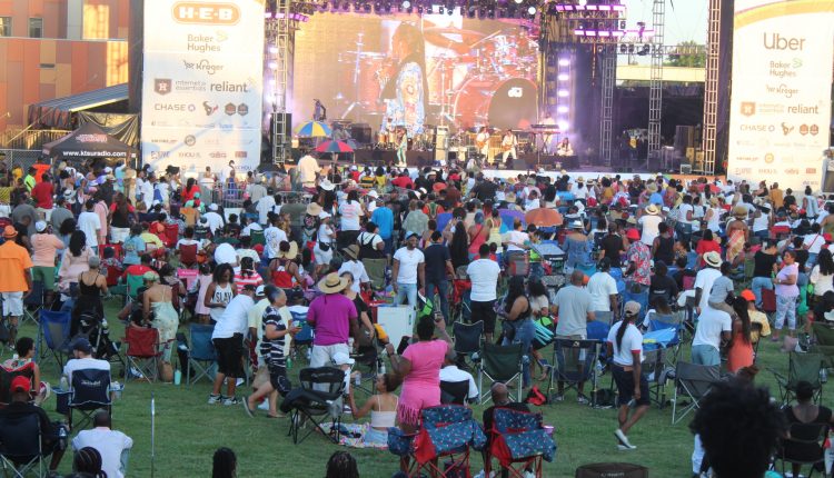 A closer shot of the stage where Sheila E. is visible on the back led screen as she performs. To the left, the black vip tent can be seen for front setting. Sheila E is in blue and her band wears white. Sponsor banners flank the stage in white with logos from numerous companies on them. People are spread out across the grass as they listen to the performance. Photograph by The Signal reporter Jared Cadore.