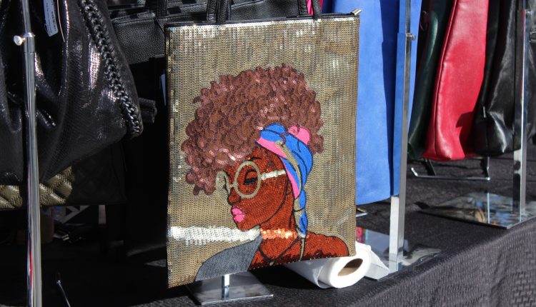 A custom purse design. The purse itself is gold. On the bag is the face of a young black woman. She wears a blue and pink band. She has pink lipstick, gold glasses, and hair in curls. Photograph by The Signal reporter Jared Cadore.