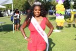 Ms. Black Houston wears a white sash with her title written on it. She wears a red dress with a silver crown on her head. Hands on her hips, she poses center for the picture on the outside grass area of the park. Ballons are in the background to her right and some park attendees to her left. Photograph by The Signal reporter Jared Cadore.