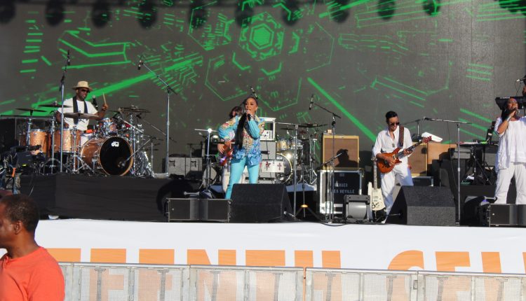 Sheila is in the center of the picture mic in her left hand as she sings wearing sky blue pants and a blue jacket. To her left is her drummer and two her right are her guitarist and a backup singer. All members of the band are wearing white. The background is a led screen showing rotating graphics. The current background is solid black with green lights in the form of a hexagon spinning around. Photograph by The Signal reporter Jared Cadore.