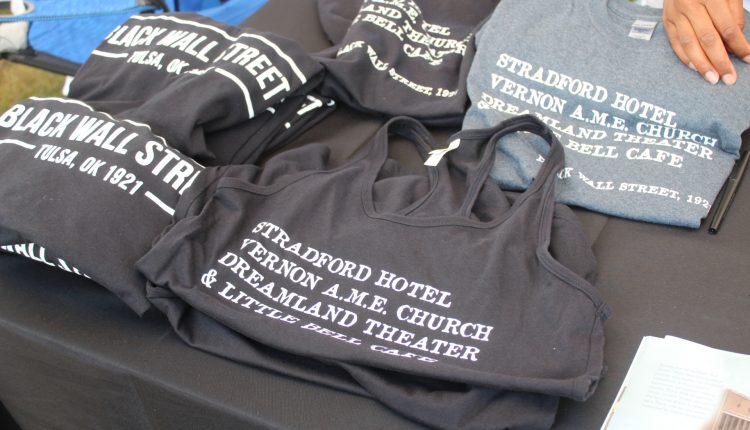 Two shirt designs sit upon this table with a black drape over it. Two of the left say "BLACK WALL STREET" with "Tulsa, OK 1921 written underneath. A white line border surrounds the text and connects to the last word line line on either side. Second shirt design says "STRADFORD HOTEL, VERNON A.M.E. CHURCH, DREAMLAND THEAER, & LITTLE BELL CAFE". Photograph by The Signal reporter Jared Cadore.