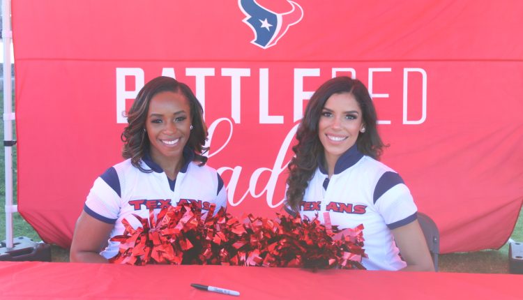 Two cheerleaders are wearing white jumpsuits with the word Texans across their chest. They each have a pair of red bombs. They're seated in front of a red background that has the Texans bull logo on it with "BATTLERED" underneath. The table they sit in front of has some pens and calendars that they sign for attendees. Photograph by The Signal reporter Jared Cadore.