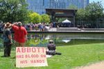 PHOTO: A group of protesters look at the stage from across a pond. They rest their sign against a cooler to prop it up. The sign reads “Abbott has blood on his hands” Photograph by The Signal Managing Editor Cesar Cardenas.