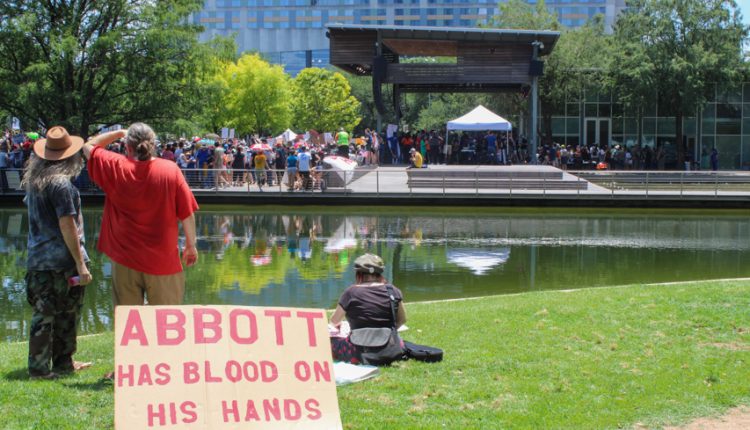PHOTO: A group of protesters look at the stage from across a pond. They rest their sign against a cooler to prop it up. The sign reads “Abbott has blood on his hands” Photograph by The Signal Managing Editor Cesar Cardenas.