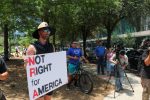 PHOTO: A protester holds a sign that reads “ Not Right for America” with the letter N,R and A in red. Two people with cameras are setting their video cameras up. Photograph by The Signal Managing Editor Cesar Cardenas.
