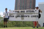 PHOTO: Two protester hold a large sign up that reads “The Big Steal” Our Children's Lives” The sign has a large faded assault riffle painted behind the letters. Photograph by The Signal Managing Editor Cesar Cardenas.