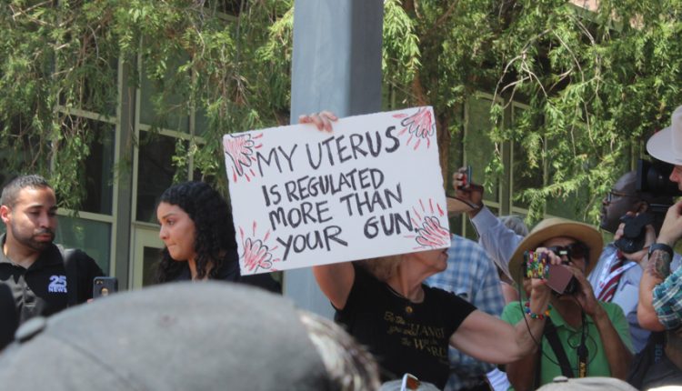 PHOTO: A protester holds a sign that reads “My uterus is more regulated than your gun” as she records the various speakers at the protest. A group of reporters stand around them. Photograph by The Signal Managing Editor Cesar Cardenas.