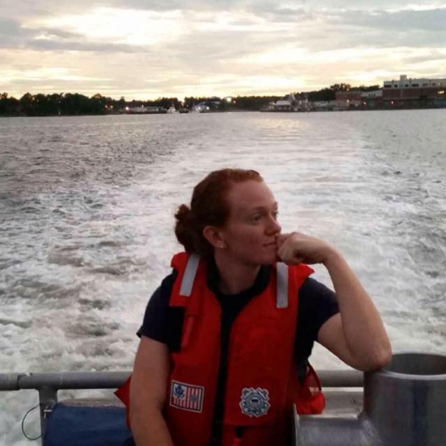 PHOTO: Coast Guard member in Coast Guard life jacket in a relaxed pose on the back of a small boat with sunset behind her.
