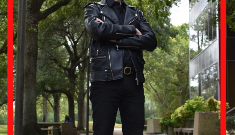 Carlos Mazziotta, Undergrad in Sociology, standing outside the Patio Café. The trees curving above him while light peaks through the trees. Dressed in an all black ensemble of a button up, slacks, and leather boots and a jacket.