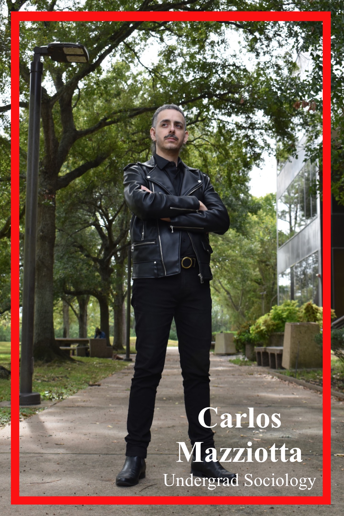 Carlos Mazziotta, Undergrad in Sociology, standing outside the Patio Café. The trees curving above him while light peaks through the trees. Dressed in an all black ensemble of a button up, slacks, and leather boots and a jacket.