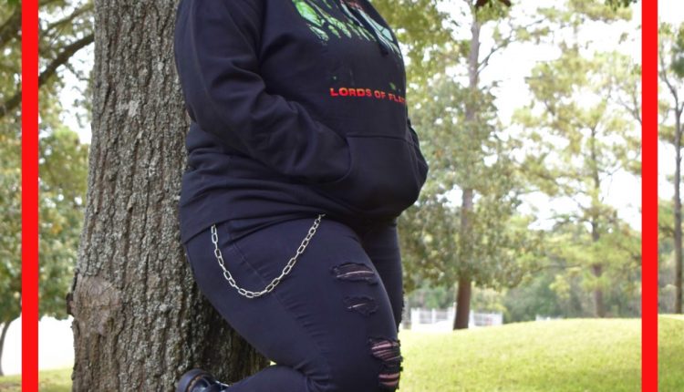 Vinnie Hughes, Undergrad in Biology, resting against a tree outside of Bayou Building. Dressed in all black attire with a graphic hoodie, ripped jeans and chain, boots, and a graphic mask to complete the ensemble.