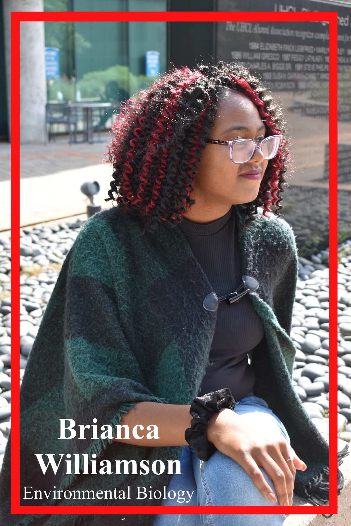 Brianca Williamson, Environmental Biology Major, sitting outside of SSCB. Sporting a black turtleneck an dark-green checkered shawl, surrounded by rocks.