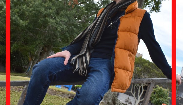 Mason Taylor, History BA Major, reclining on a stair rail. Dressed in matching blue long-sleeved shirt and jeans, he uses a pumpkin orange vest-parka and aquamarine shoes to add pops of color, completing the loo with a dark sepia striped scarf.