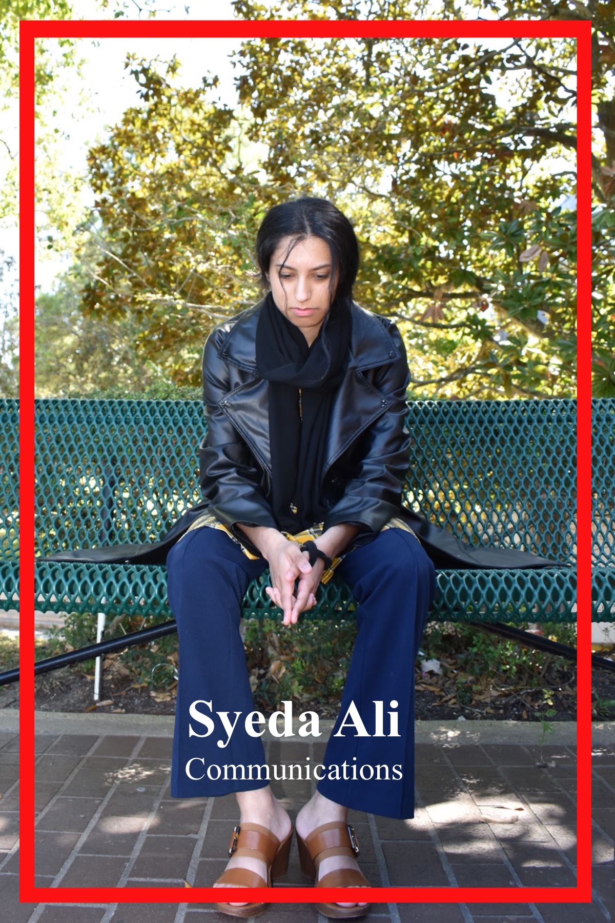Syeda Ali, Communications major, sitting on bench in front of tree foliage. Wearing a long yellow gingham blouse, she pairs it with navy blue flared slacks and brown buckled sandals. Atop she covers up with a black leather trench coat, and a black scarf.