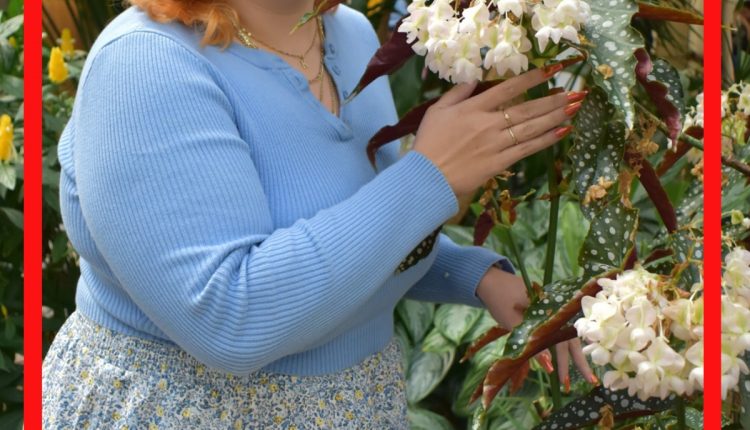 Jessi Cooley, Financial Aid Counselor and Instruction Design and Tech major, cupping white flowers in the Bayou Building botanical garden. She wears a white minidress with blue and yellow floral decals, and wears a complementing baby blue cropped cardigan to complete the look.