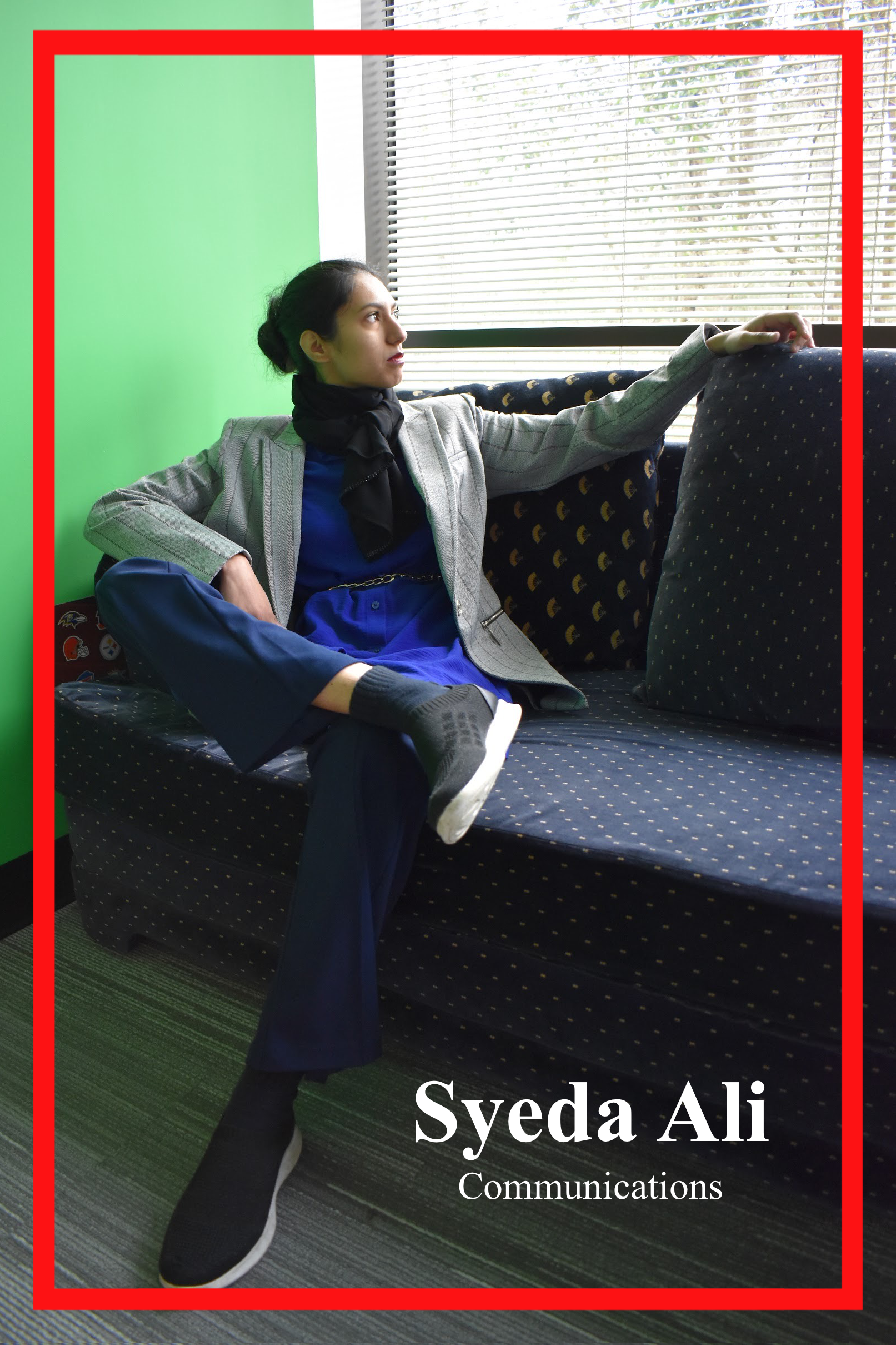 Syeda Ali, Communications Major, lounging on a blue couch. Wearing a minimal grey pin-stripe blazer with a strong shoulder atop a chain-belted azure blue dress with navy pants. A black scarf tied adoringly around her neck.